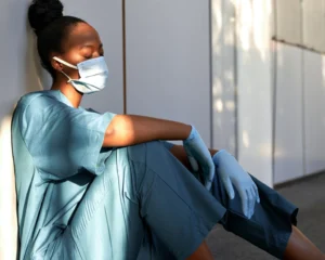 an exhausted medical professional wearing a mask and gloves napping seated against the wall in a hallway