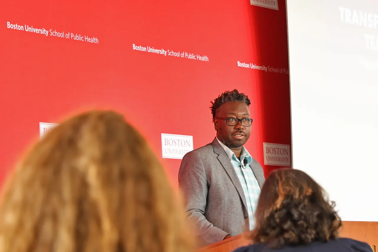 a man speaking at a lectern in front of a Boston University backdrop in a room full of people