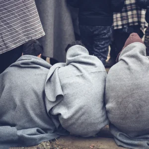 Three people sitting on the ground wrapped in gray blankets with people standing in front of them.