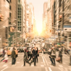 A blurred image of city life, with people walking a crosswalk and cars moving in the background